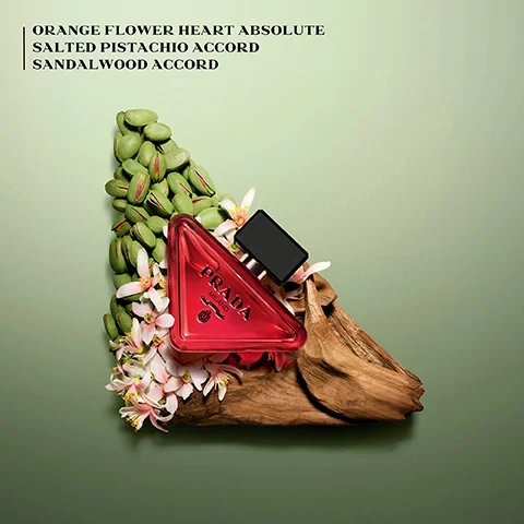 Image 1 - A red triangle perfume bottle sits on driftwood with pistachios and flowers, with text ORANGE FLOWER HEART ABSOLUTE SALTED PISTACHIO ACCORD SANDALWOOD ACCORD PRADA MILANO DAL 1913.
